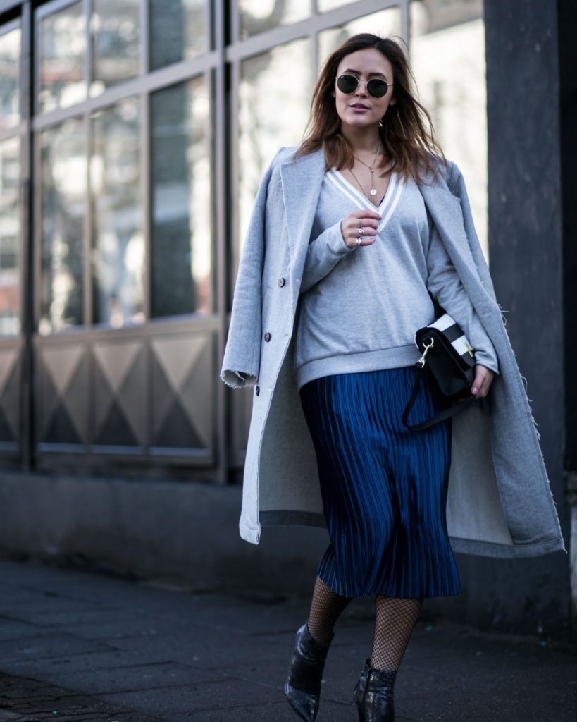 langer Wollmantel in hellblau, hohe schwarze spitze Stiefeletten in Schlangenleder Optik, mittellangen blauen Plissee Rock, lässiges Sweatshirt mit tiefem V-Ausschnitt